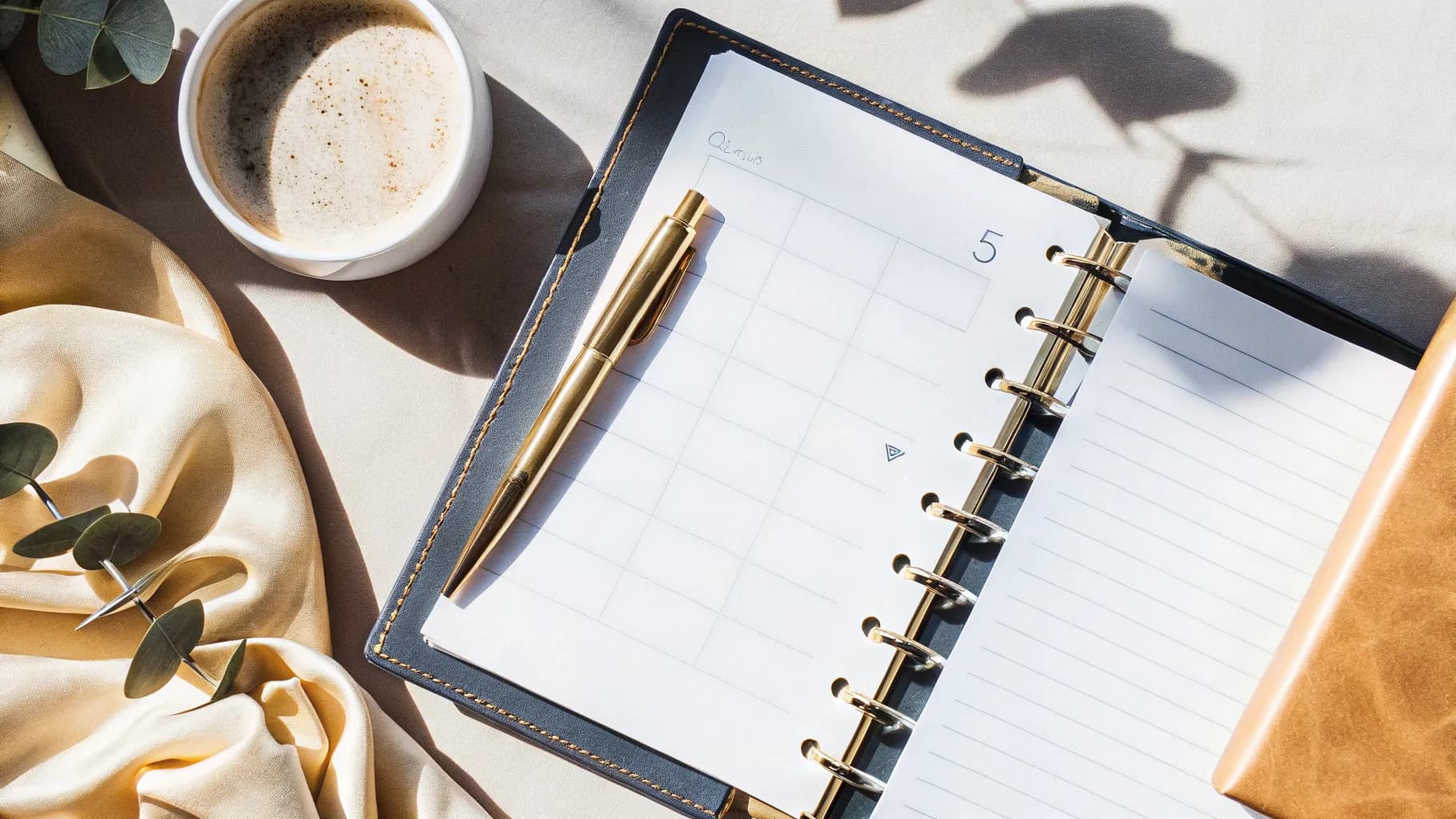 An open wedding planner with a checklist, a gold pen, a sprig of eucalyptus and a cup of coffee on a soft, sunlit table