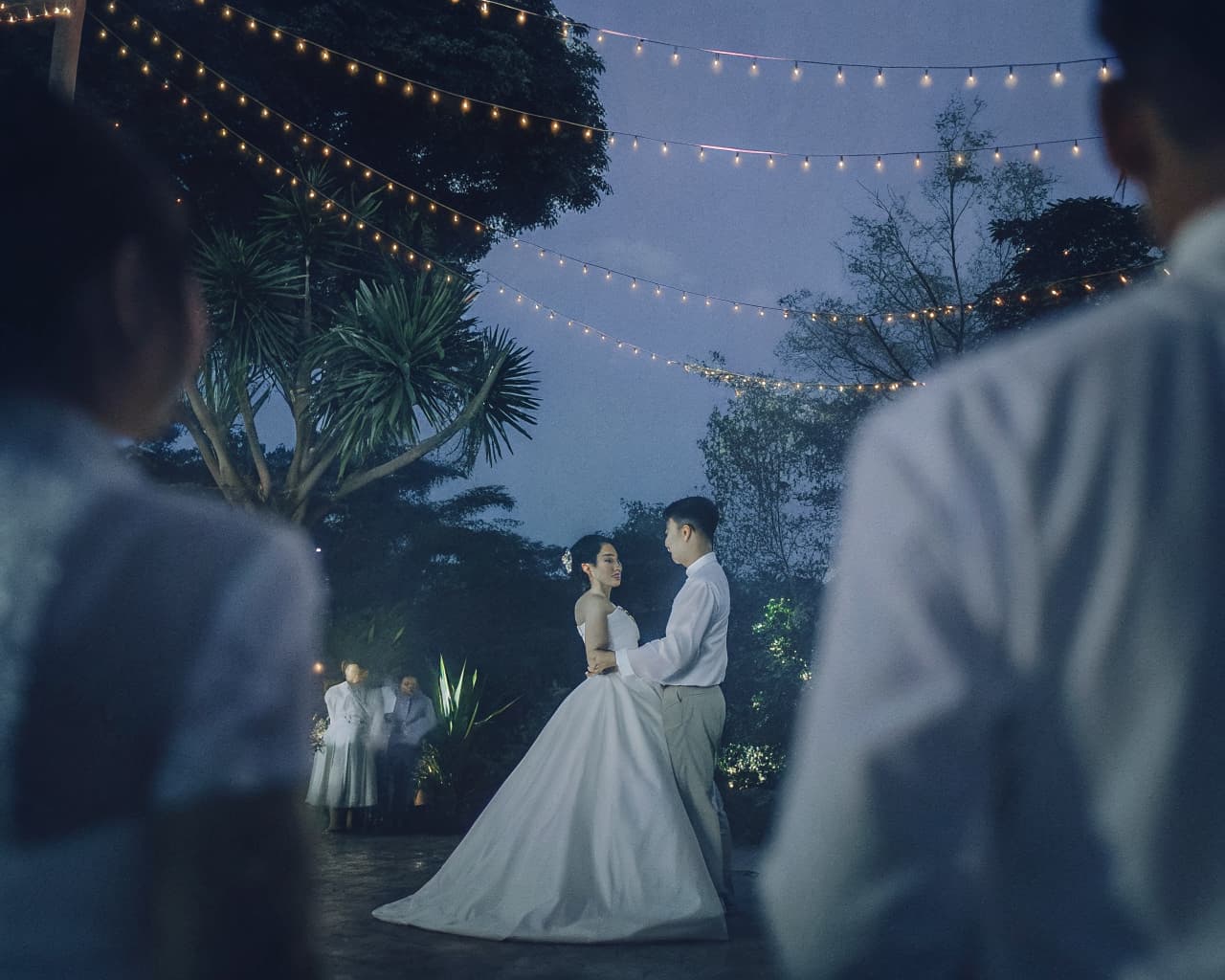 A couple dancing under strings of lights at dusk, seen between two guests